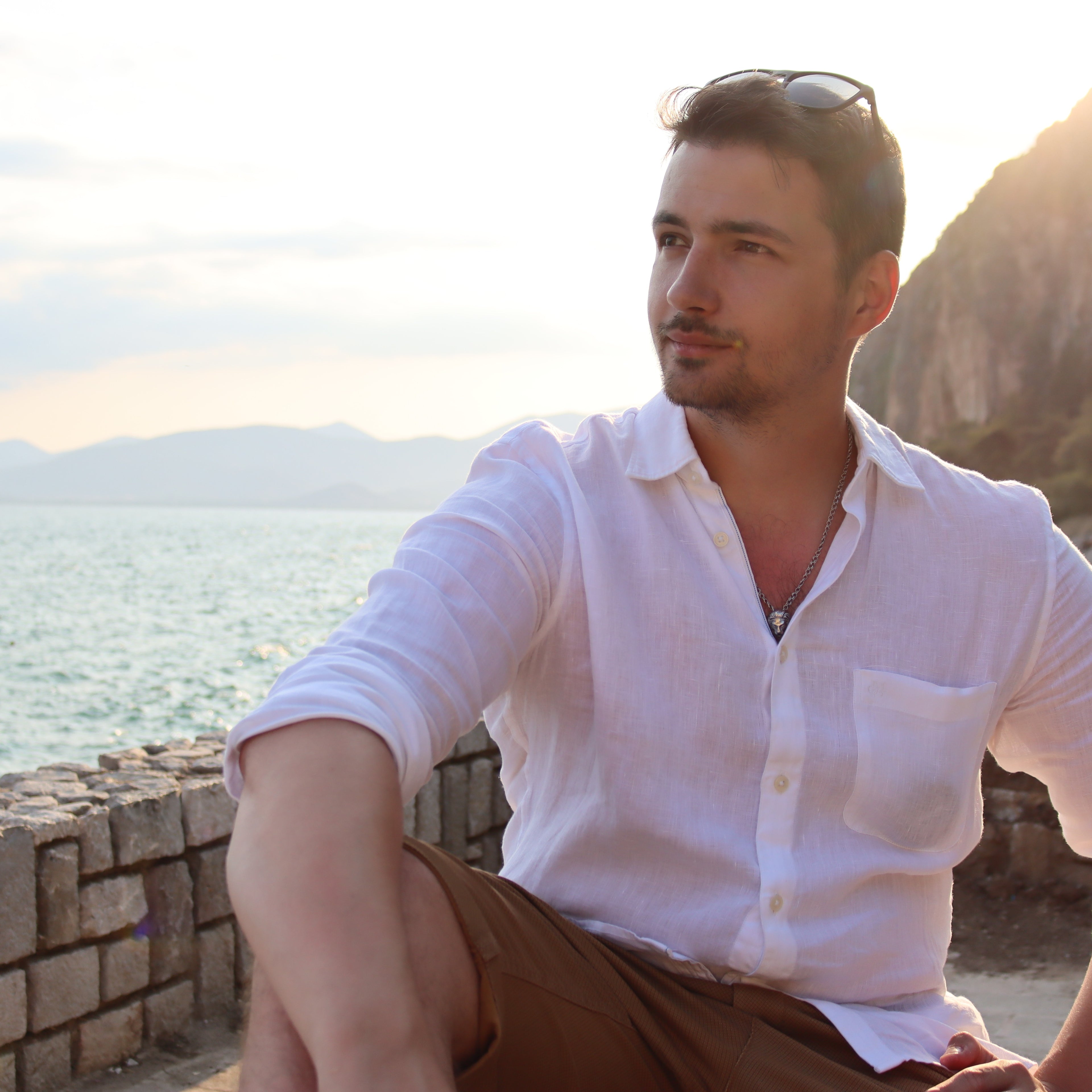 Photo of Konstantinos Giouzakov, one of the founders of Boardom Games, sitting by the sea in a white shirt for the About Us section of the party game brand website.